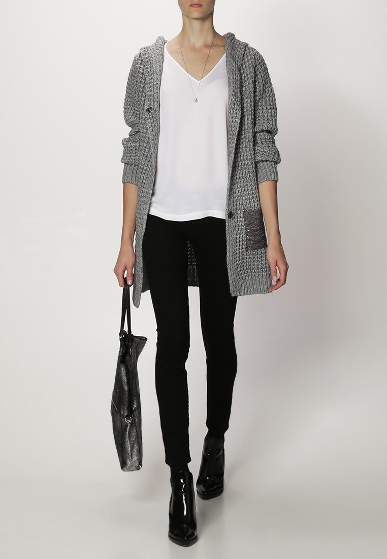 Cardigan grigio lavorato a maglia con cappuccio, top bianco a scollo a V, pantaloni neri skinny e stivaletti neri lucidi, mentre tiene una borsa tote nera.
