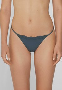 Femme portant un slip en dentelle teint émeraude foncé, se tenant avec les bras détendus sur les côtés contre un fond neutre.