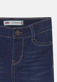 Tmavě modré džíny z denimu s velmi úzkým střihem, se zavíráním na knoflíky a kontrastním prošitím podél kapes a švů.