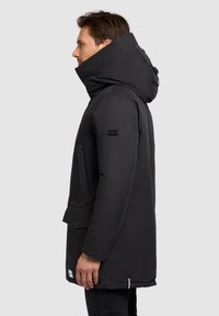 Muž ve černém kabátu s kapucí stojící vlevo, opřený o jednolitý světlý pozadí.