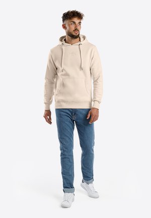 Man with curly hair wearing a beige hoodie, blue jeans, and white sneakers standing against a plain light background.