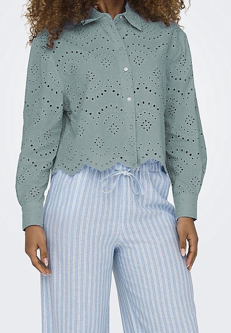 Femme portant un chemisier vert à manches longues en dentelle ajourée et un pantalon bleu clair à cordon avec des rayures verticales blanches.
