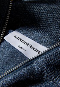 Närbild av blått stickat tyg med en silverfärgad dragkedja som är delvis öppen och avslöjar en vit etikett med texten "LINDBERGH SLIM FIT."