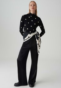 Haut à manches longues noir à pois blancs, associé à un pantalon noir ample ; une écharpe rayée noire et blanche nouée autour de la taille.