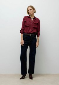Modelka stojí na neutrálním pozadí v bordó košili, tmavých džínách a stylových podpatcích, ukazující moderní outfit.