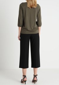 Vrouw staat met haar rug naar de camera, gekleed in een olijfgroene top met driekwartmouwen, zwarte cropped broek en zwarte hoge hakken.