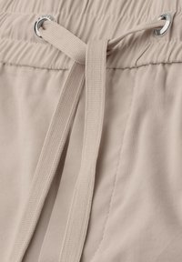 Ceinture en tissu beige avec un élastique froncé et un cordon plat tissé passé à travers des œillets métalliques, noué d'un simple nœud.