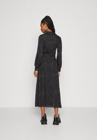 Zwarte maxi-jurk met stippen, voorzien van lange mouwen, een cinched taille en een luchtige rok. Draagt met dikke zwarte schoenen, zicht vanuit de achterkant.