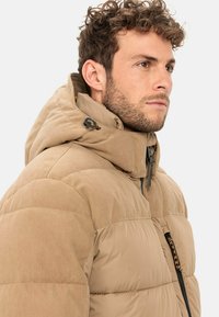 Beige Daunenjacke mit einer Cordstrick-Textur und hohem Kragen. Sie verfügt über eine Kapuze, einen Reißverschluss und eine Akzenttasche mit einem gemusterten Detail.