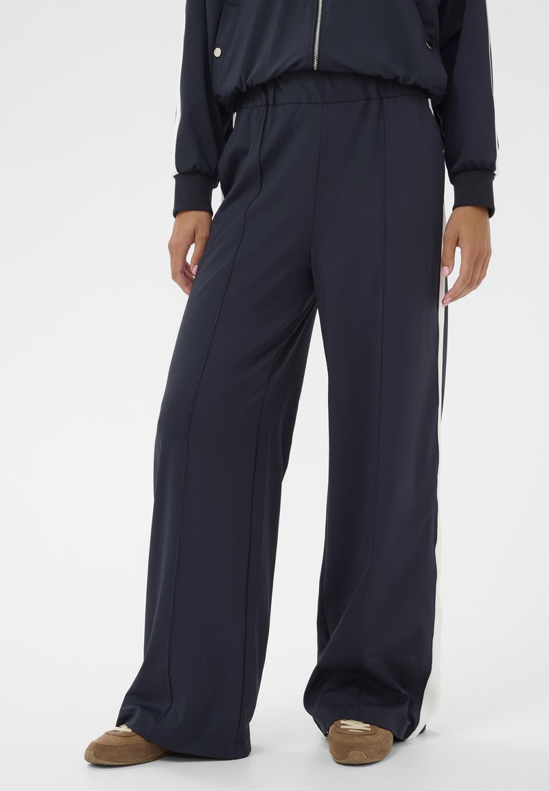 Personne portant un pantalon large bleu marine avec des bandes blanches sur le côté et des chaussures décontractées en daim marron, se tenant sur un fond blanc uni.