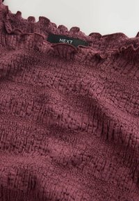 Tessuto bordeaux testurizzato con smock elastico e bordo arricciato, con etichetta nera con la scritta "NEXT" in bianco.