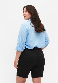 Camisa de lino azul claro con mangas enrolladas, combinada con pantalones cortos de mezclilla negros de talle alto que cuentan con bolsillos traseros y un acabado suave.