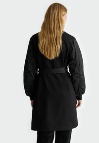 Čierny kabát s dlhými, nadýchanými rukávmi z kontrastného materiálu. Má opasok v páse a hladkú textúru. Jednoduchý, elegantný dizajn.