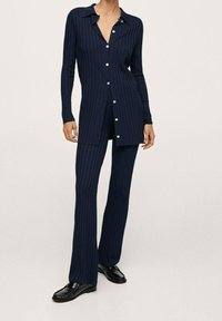 Žena oblečená v námořnické modré, žebrované tunice s dlouhým rukávem a knoflíky, s odpovídajícími rozšířenými kalhotami a černými koženými mokasínami, stojící na jednolitém pozadí.