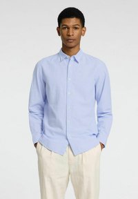 Camicia blu chiaro a maniche lunghe con una texture morbida, colletto e patta frontale, abbinata a pantaloni color crema.