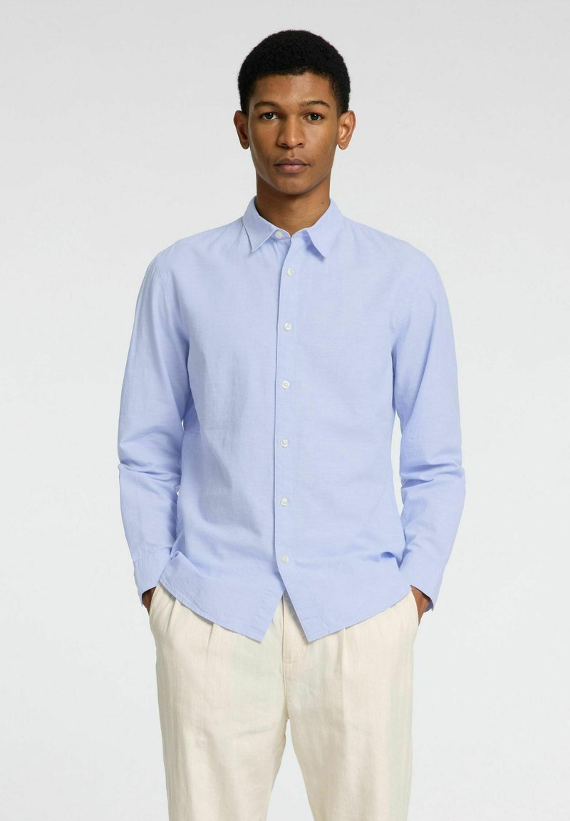 Camicia blu chiaro a maniche lunghe con una texture morbida, colletto e patta frontale, abbinata a pantaloni color crema.