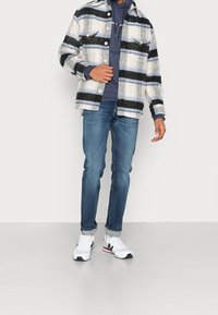 Chemise en flanelle à motif carreaux bleu, noir et blanc, associée à un sweat à capuche sombre, un jean bleu, des baskets blanches et des manches retroussées.