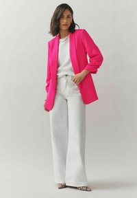 Fuchsiové sako so štruktúrovanými ramenami, doplnené bielym topom a širokými bielymi nohavicami. Celkový vzhľad dopĺňajú členkové podpätky s hadím vzorom.