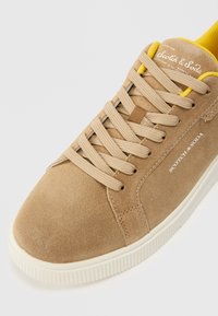 Zapatillas de gamuza marrón con cordones beige, detalles amarillos y suela de goma blanca. Presenta un estampado de logo en el costado y un upper texturizado.