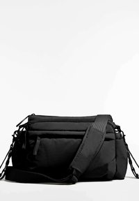Črno oblazinjen torbica čez ramo z dvema žepoma z zadrgo, nastavljivim trakom in teksturirano površino. Ima vezalke in elegantno zasnovo.
