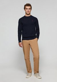 Marinblå kabelstickad tröja med rund halsringning, i kombination med khakifärgade chinos och ljusa sneakers. Enkelt mönster med ribbade muddar och nederkant.