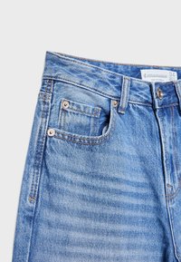 Vaaleansinisiä farkkuja, joissa on korkea vyötärö, viisi taskua, kuparinvärisiä nappiyksityiskohtia ja hienovaraisia kulutustekijöitä kankaassa.