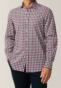 Mann mit einem langärmeligen Hemd mit rot-blau-weißer kariertem Muster und dunkler Hose, die Hände teilweise in den Taschen.