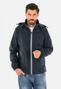 Giacca impermeabile blu navy con due tasche frontali, dettagli in cerniera bianca, cappuccio regolabile e polsini elastici, indossata sopra una camicia bianca.