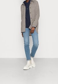 Rutigt långskjortjacka i beige och brunt, lager på lager över en marinblå huvtröja. Ljusblå jeans och vita kraftiga anklar stövlar kompletterar outfiten.