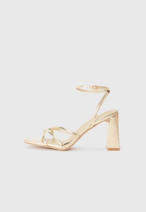 Zlaté sandále na vysokom podpätku so štvorcovou špičkou, prekríženými remienkami a remienkom okolo členka. Lesklý povrch a blokový podpätok zabezpečujú stabilitu.