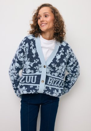 Jonge vrouw die glimlacht, gekleed in een blauw-wit vest met bloemenpatroon en de tekst "ZUU BIZU" erop, en een donkerblauwe broek, tegen een effen achtergrond.