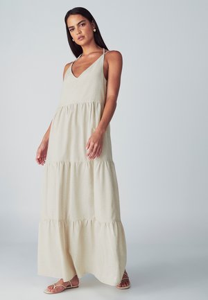 Žena s tmavými vlasy, oblečená v bezramínkových béžových vrstvených maxi šatech a plochých sandálech, stojící před jednobarevným světlým pozadím.