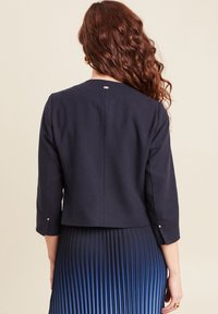 Giacca cropped navy con maniche a tre quarti, tessuto testurizzato e bottoni argentati sugli polsini, abbinata a una gonna plissettata a gradiente.