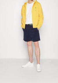Gul zip-up hoodie över en vit t-shirt, tillsammans med marinblå shorts och vita sneakers; slät textur och avslappnad design.