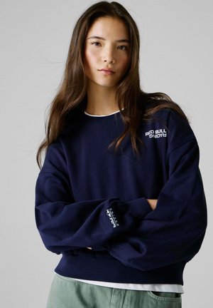 Granatowa bluza z długimi, oversize rękawami. Na przodzie i mankiecie znajduje się logo "Red Bull Racing". Miękka tkanina, luźny krój.