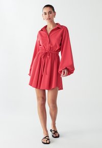 Rotes Hemdblusenkleid, tailliert, mit langen Puffärmeln, aus leichtem Stoff, mit ausgestelltem Rock, kombiniert mit schwarzen Sandalen.