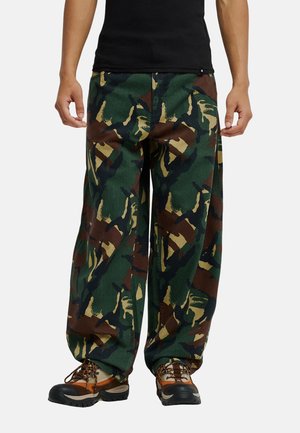Persoon met een groene, bruine en zwarte camouflagebroek, bruine wandelschoenen en een zwart shirt, rechtop staand op een witte achtergrond.