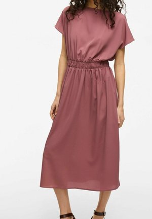 Kobieta ubrana w różową, bezrękawną sukienkę midi z marszczoną talią i okrągłym dekoltem, w zestawie z ciemnobrązowymi sandałami na kostkę z paskiem.