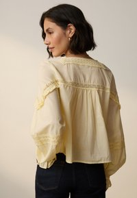 Żółta bluzka z koronkową lamówką, falbaniastymi rękawami i luźnym krojem. Miękki materiał o lekkiej fakturze oraz szeroki, luźno opadający dekolt.