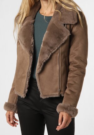 Vrouw draagt een bruine suède jas met grote bontkraag en manchetten, gecombineerd met een donkergroene top en een zwarte broek tegen een effen achtergrond.