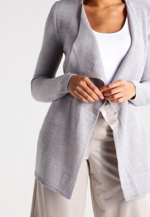 Cardigan en tricot gris clair avec un design texturé, ouvert sur le devant, manches longues et détails côtelés aux poignets et à l'ourlet. Associé à un haut blanc.