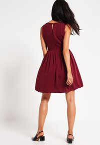 Burgunderfarbenes ärmelloses Kleid mit enger Passform am Oberteil und gefälteltem Rock. Es hat einen Schlüsselrücken und einen glatten, strukturierten Stoff.