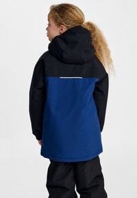 Veste imperméable avec une partie inférieure bleue et une partie supérieure noire, dotée d'une capuche. Texture lisse et accent réfléchissant au dos.