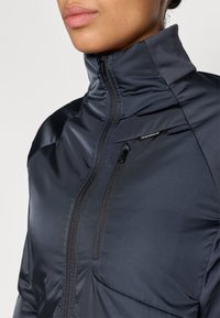 Veste matelassée navy avec col haut, fermeture éclair à l'avant, poches latérales et texture lisse. Présente un logo sur la poitrine et un design ajusté.