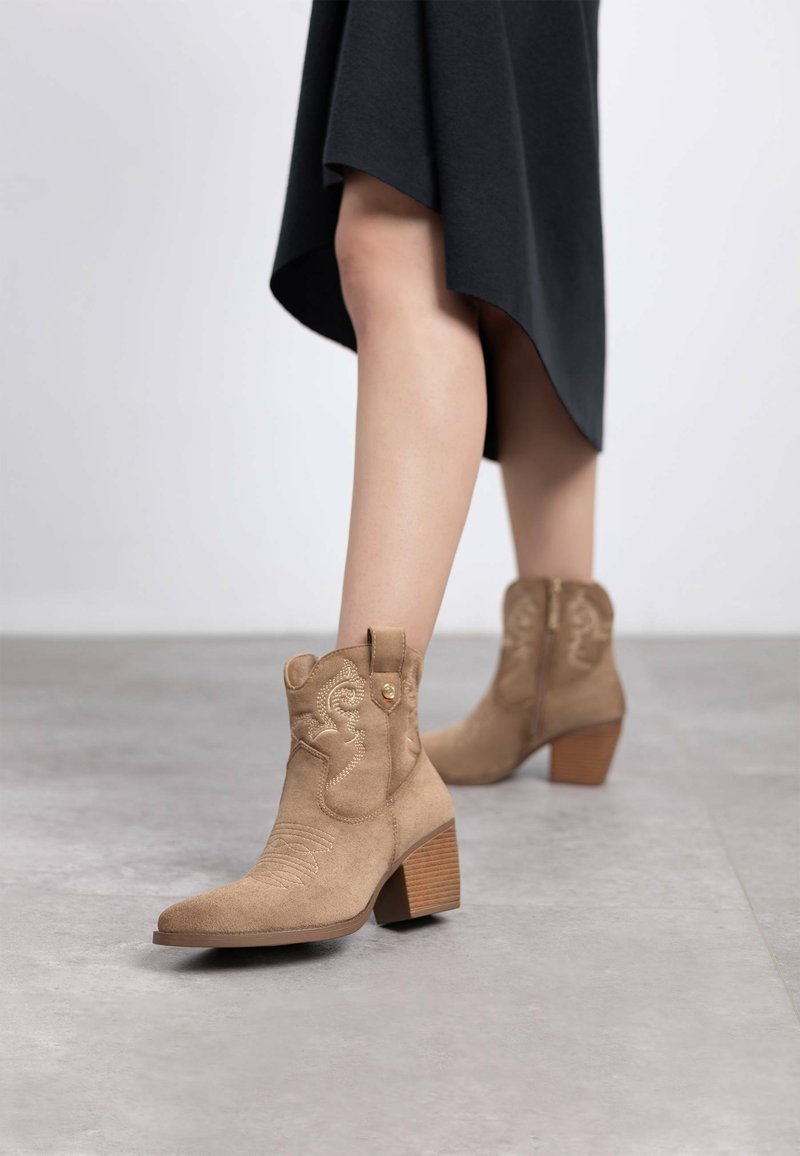 Botines de ante beige con puntera ligeramente afilada, tacón de madera apilada, costuras decorativas y cierre de cremallera lateral.