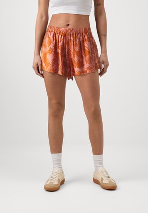 Pantalones cortos naranjas y rosas con un patrón de hojas tropicales, que cuentan con una cinturilla elástica y un corte relajado. Combinados con calcetines blancos y zapatillas beige.