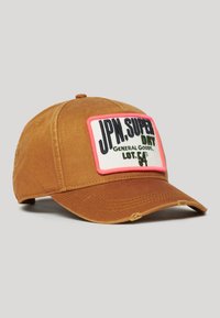 Gorra de algodón marrón con visera curva, que presenta una placa rectangular en rosa y blanco con texto negro. Acabados desgastados en la visera.