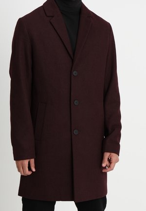 Ciemnobrązowy wełniany płaszcz z trzema czarnymi guzikami, klapami z wycięciem i bocznymi kieszeniami, noszony na czarnym golfie.