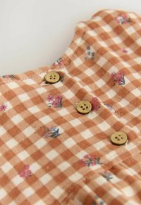 Tejido marrón con cuadros y estampados florales, con tres botones de color claro en el escote. Textura suave y diseño casual.