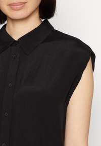 Svart ärmlös skjorta med knäppning fram, mjuk textur, spetsig krage och subtil glans; har detaljering vid axelsömmarna.